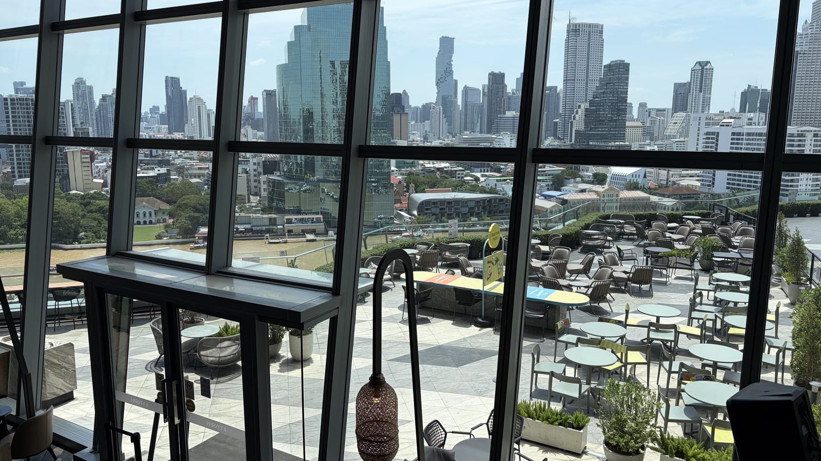 窓越しに見た屋外テラス。市松模様の床にテーブルとチェアが並び、奥に対岸のビル群がそびえる