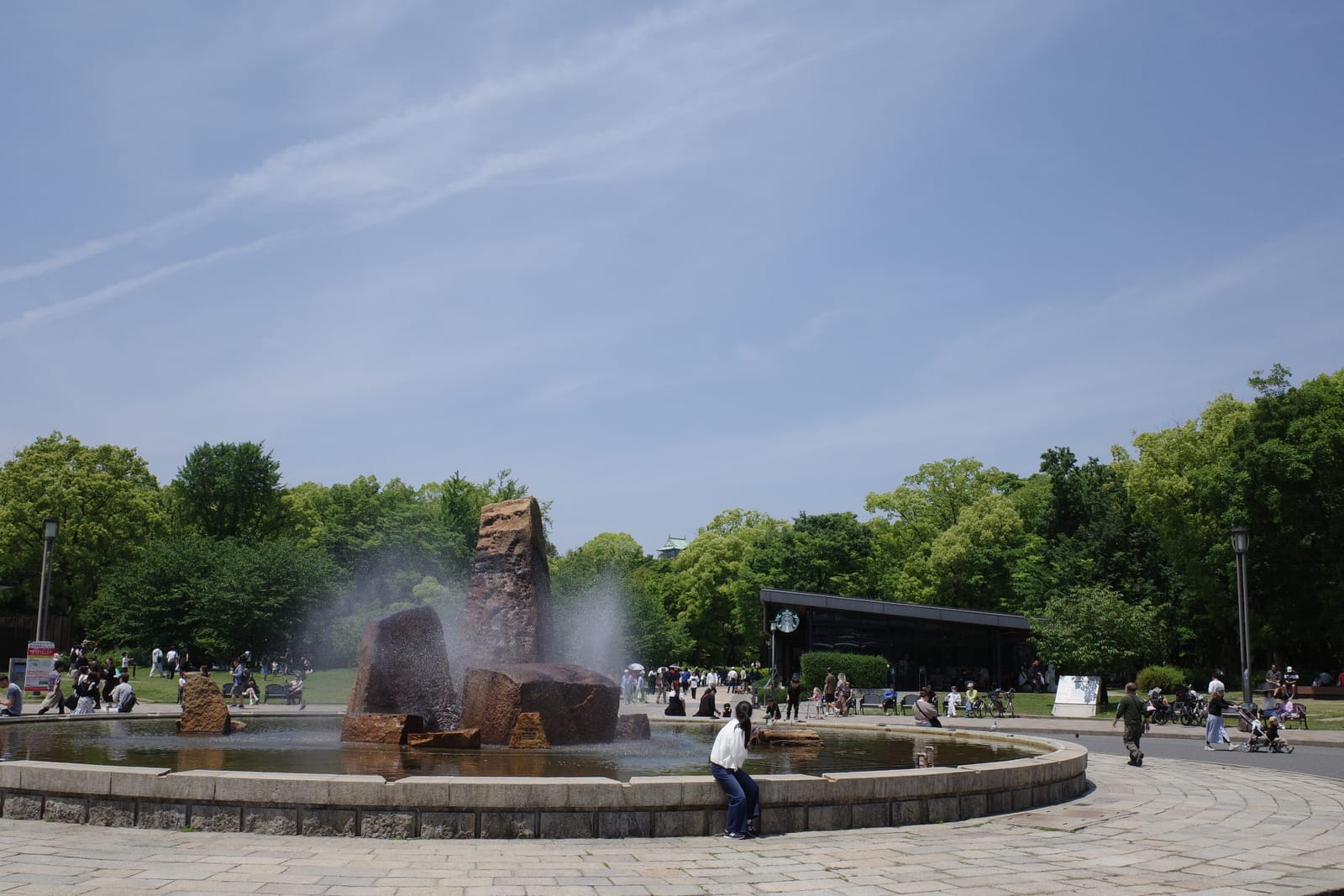 大阪城公園の噴水広場越しに見える大阪城公園森ノ宮店。屋根の低い黒い店舗と緑の森、奥にうっすら大阪城の天守閣が見える