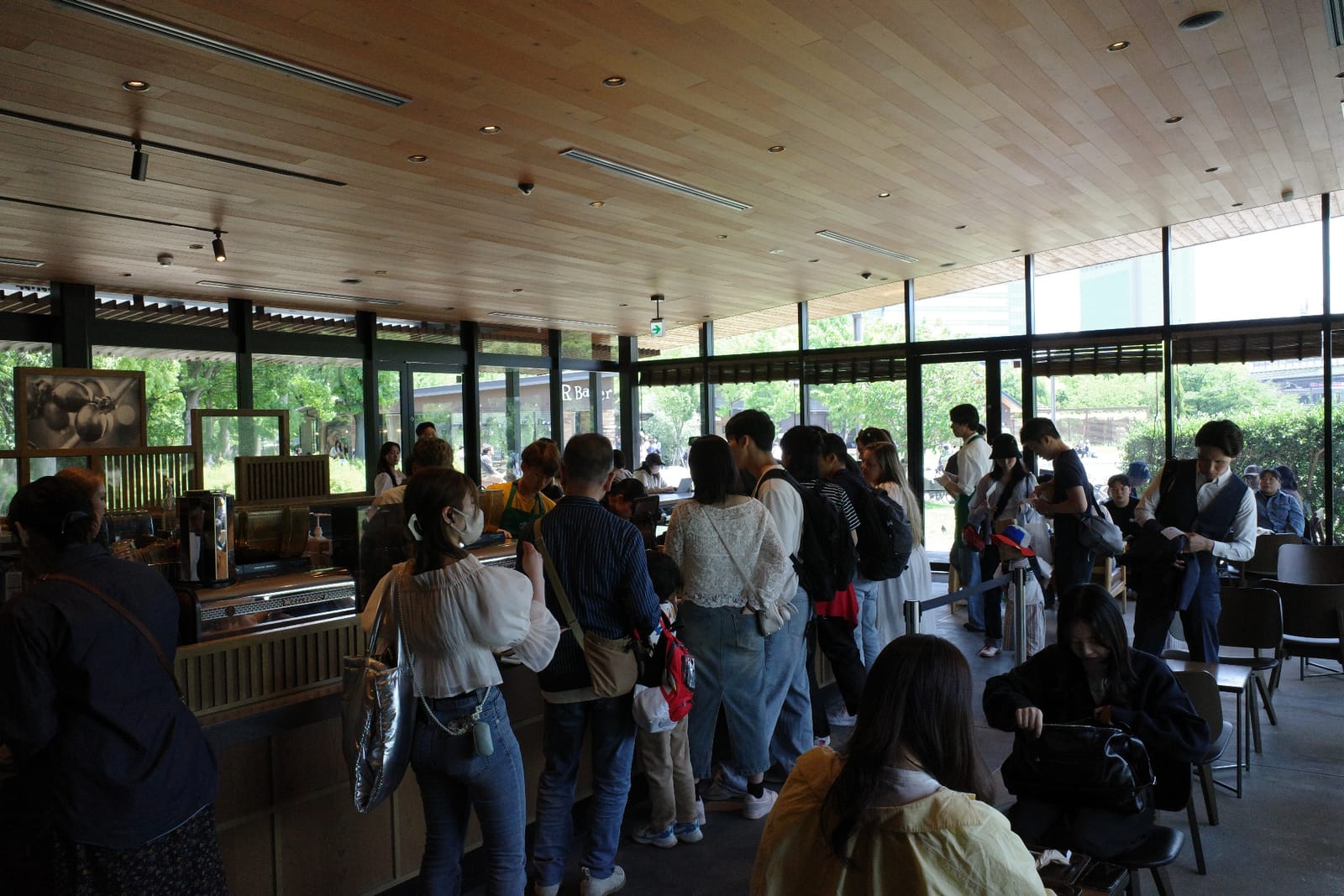 店内の混雑シーン。木の板張りの天井とダウンライト、ガラスで囲まれた壁の向こうに公園の緑、レジ前に並ぶ長い行列