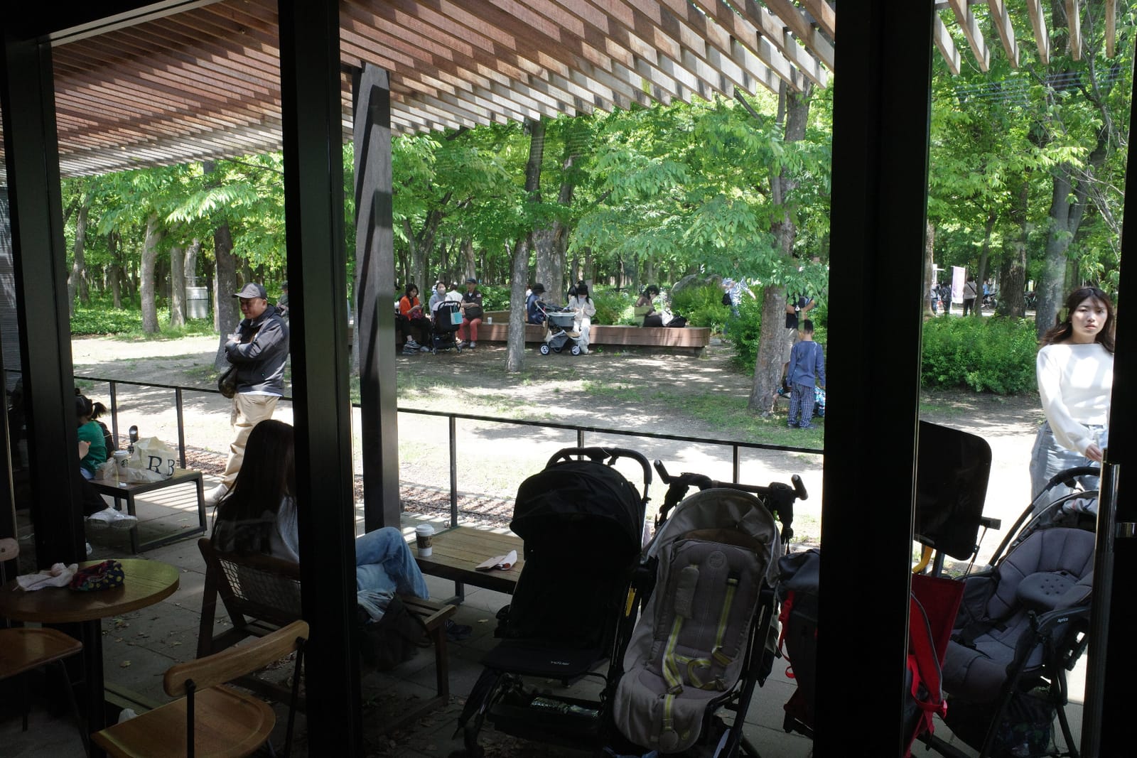 店内から見た店外。手前にベビーカーが並び、ガラスの向こうの公園では木陰のベンチに人々が座って休んでいる