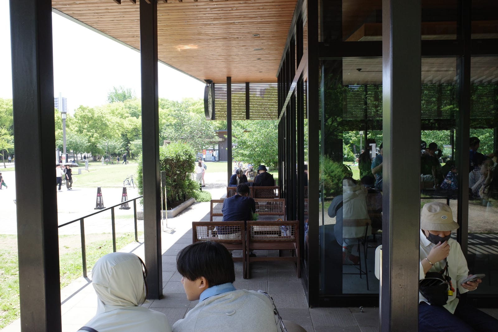 屋根付きテラス席。木の梁の天井、籐っぽい木のイスとテーブル、ガラス越しに店内が見え、奥には公園の緑が広がる