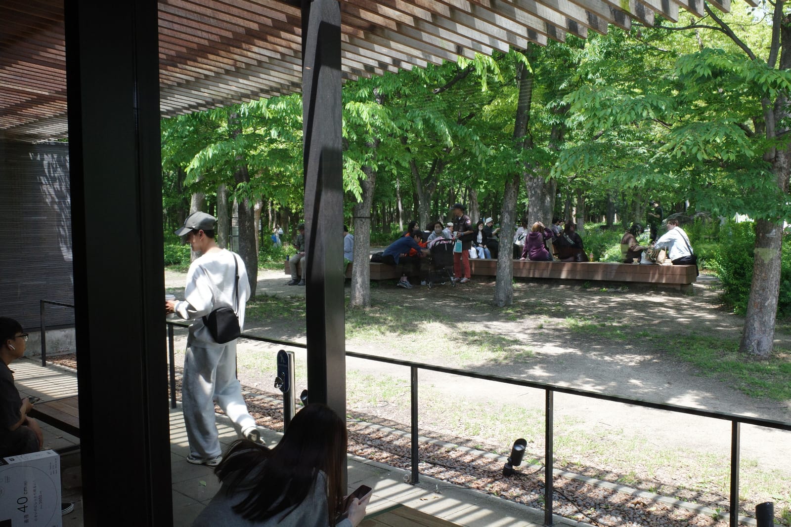 テラスの外、公園側のベンチに腰掛けてくつろぐ人々。木陰と緑、コの字型に並ぶベンチ