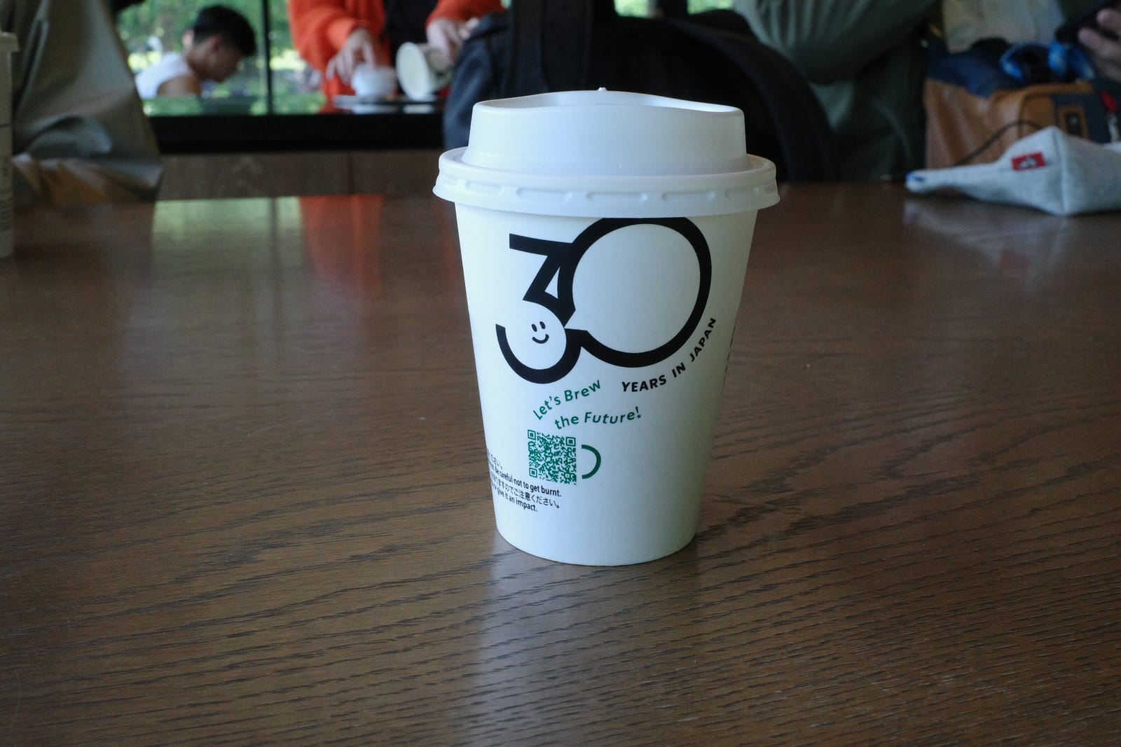 木目のテーブルに置かれた紙カップ。スターバックス日本上陸 30 周年ロゴの「Let's Brew the Future!」が描かれている。背景には公園の緑とガラス越しの席
